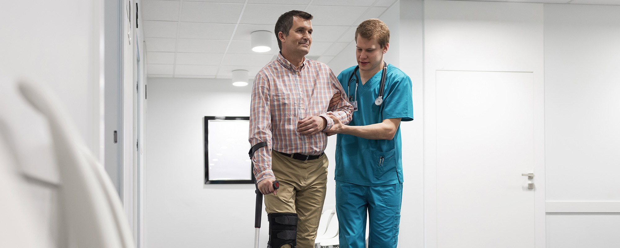 nurse assisting patient with injured leg in walking at hospital corridor